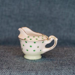 May include: A small, white creamer with green polka dots and a gold rim. The handle is also gold. The creamer sits on a small, round base. The creamer is likely made of porcelain or ceramic.