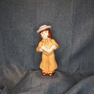 May include: A ceramic figurine of a girl wearing a light blue hat and a brown dress. She is holding a book open in her hands. The figurine is painted in shades of brown, white, and blue.