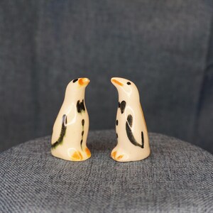 May include: Two ceramic penguin figurines. The penguins have a cream-colored body with black spots and markings. They have orange beaks and feet. The figurines are standing on a gray surface.