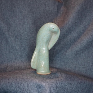 May include: A ceramic figurine of a bird with a speckled light blue glaze. The bird is standing with its head bent down.