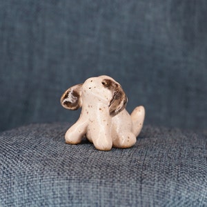 May include: A small ceramic sculpture of a dog with brown ears and a white body. The dog is sitting up with its head tilted up.