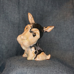 May include: A ceramic figurine of a dog with black and white fur. The dog has a large, expressive face and is sitting with its head tilted to the side.