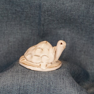 May include: A white ceramic turtle figurine with a detailed shell and a slightly upturned head. The turtle is sitting on a plain background.