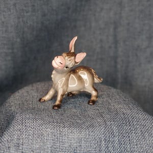 May include: A small ceramic donkey figurine with a glossy finish. The donkey is colored in shades of brown and gray with a light pink muzzle and inner ears. It is depicted in a standing position with its head tilted upward and eyes closed.