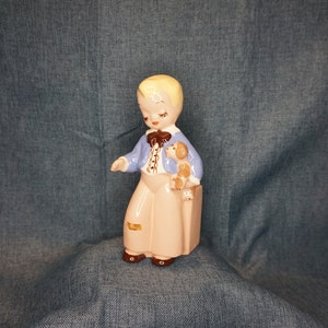 May include: A ceramic figurine of a boy with blonde hair wearing a blue jacket and brown pants, holding a small dog. The figurine is painted in shades of blue, brown, and white.