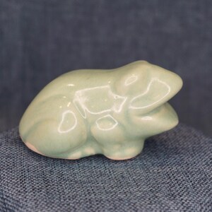 May include: A small, smooth, light green ceramic frog figurine in a seated position. The sculpture features a glossy, reflective glaze with subtle variations in color tone, highlighting the rounded contours of the frog's body and head.