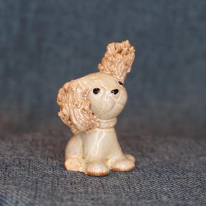 May include: A small ceramic dog figurine with a glossy beige finish. The sculpture features textured, curly ears, a smooth body, and painted black eyes and nose. The dog is depicted in a seated position with one ear standing upright and one ear down.