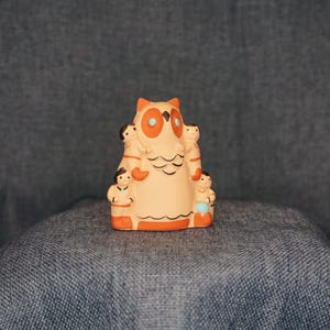 May include: A ceramic owl figurine with orange and cream coloring. The owl has large orange eyes and is surrounded by small figures. The figurine is sitting on a blue surface.