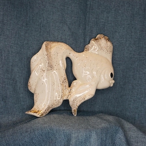 May include: A ceramic wall plaque shaped like a goldfish, featuring a glossy cream-colored finish with speckled gold accents throughout its body and flowing fins. The fish has a single visible black eye and a sculpted, textured surface design.
