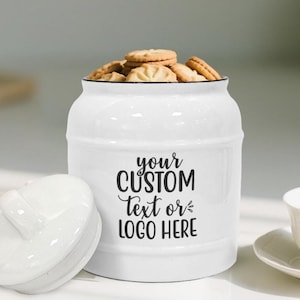 Op de afbeelding: Een witte keramische koektrommel met een zwarte rand en de tekst "your CUSTOM Text or LOGO HERE". De pot is gevuld met koekjes. Het deksel ligt ernaast.