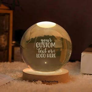 May include: A clear glass sphere lamp resting on a circular light-coloured wooden base. The sphere features the engraved text "your CUSTOM text or LOGO HERE" in the centre. The wooden base has the text "Custom Engraving" printed on the front.