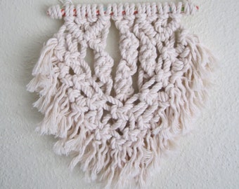 May include: A white macrame wall hanging with a fringe border. The hanging is made with a natural cotton cord and is attached to a wooden dowel.