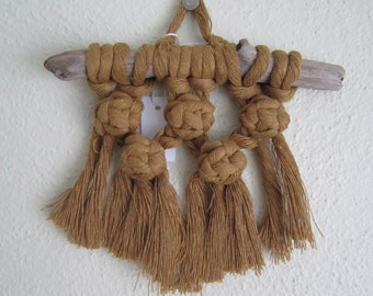 May include: A macrame wall hanging with a driftwood base and brown tassels. The wall hanging features a geometric pattern of knots.