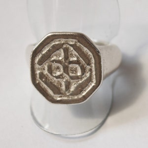 Könnte beinhalten: Ein silberner Ring mit einer achteckigen Fläche, die mit einem erhabenen, geometrischen Muster verziert ist. Der Ring wird auf einem weißen Hintergrund gezeigt.