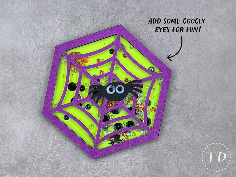 May include: A purple and green hexagon-shaped craft with a spider web design. The web is filled with glitter and googly eyes. The text "ADD SOME GOOGLY EYES FOR FUN!" is written above the craft.