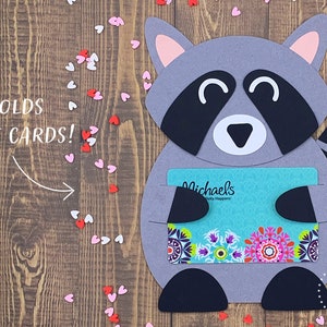 May include: A gray raccoon holding a gift card for Michael's, a craft store. The raccoon is holding the gift card in its paws and has a happy expression on its face. The gift card is turquoise blue with a colorful floral design. The background is a wooden surface with pink and white hearts scattered around.