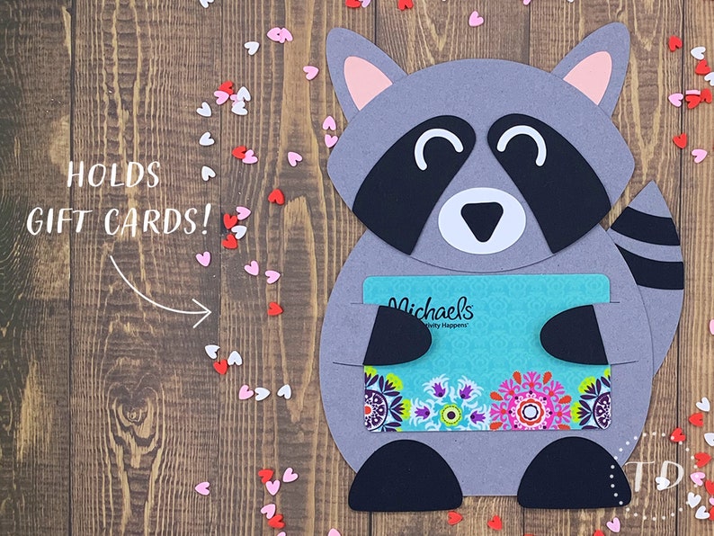 May include: A gray raccoon holding a gift card for Michael's, a craft store. The raccoon is holding the gift card in its paws and has a happy expression on its face. The gift card is turquoise blue with a colorful floral design. The background is a wooden surface with pink and white hearts scattered around.