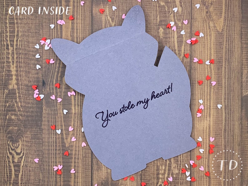 May include: A gray card shaped like a fox with the text "You stole my heart!" written on it. The card is surrounded by small pink and red hearts on a wooden background.