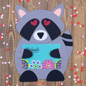 May include: A gray raccoon with pink ears and a black tail holding a gift card for Michaels, a craft store. The raccoon has red glitter hearts for eyes and is surrounded by pink and white hearts.