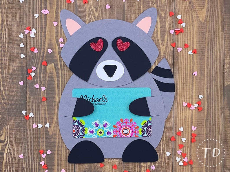 May include: A gray raccoon with pink ears and a black tail holding a gift card for Michaels, a craft store. The raccoon has red glitter hearts for eyes and is surrounded by pink and white hearts.