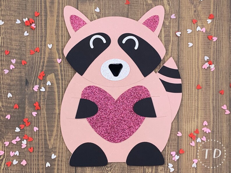 May include: A pink raccoon holding a pink glitter heart. The raccoon has black eyes, a black nose, and a black and white striped tail. The background is a wooden surface with pink and white hearts scattered around.