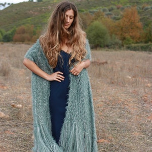 Puede incluir: Un chal largo y fluido de color verde azulado con flecos est&aacute; drapeado sobre un vestido azul marino. El chal tiene un aspecto suave y texturizado. El fondo es un campo con una colina a lo lejos.