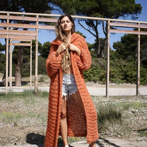 May include: A long, orange knitted cardigan with a hood. The cardigan is open at the front and has a loose fit. The model is wearing the cardigan with denim shorts and white trainers.
