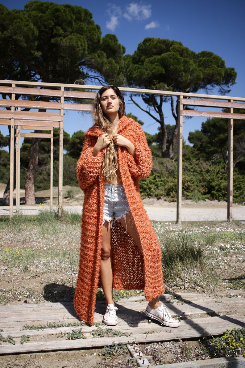 May include: A long, orange knitted cardigan with a hood. The cardigan is open at the front and has a loose fit. The model is wearing the cardigan with denim shorts and white trainers.