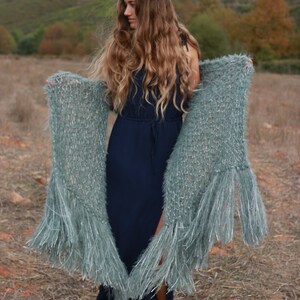 Puede incluir: Una mujer con un vestido azul marino largo y botas negras, sosteniendo un chal verde azulado claro con largas borlas. El chal est&aacute; hecho de un material suave y texturizado. El fondo presenta un campo y un cielo nublado.