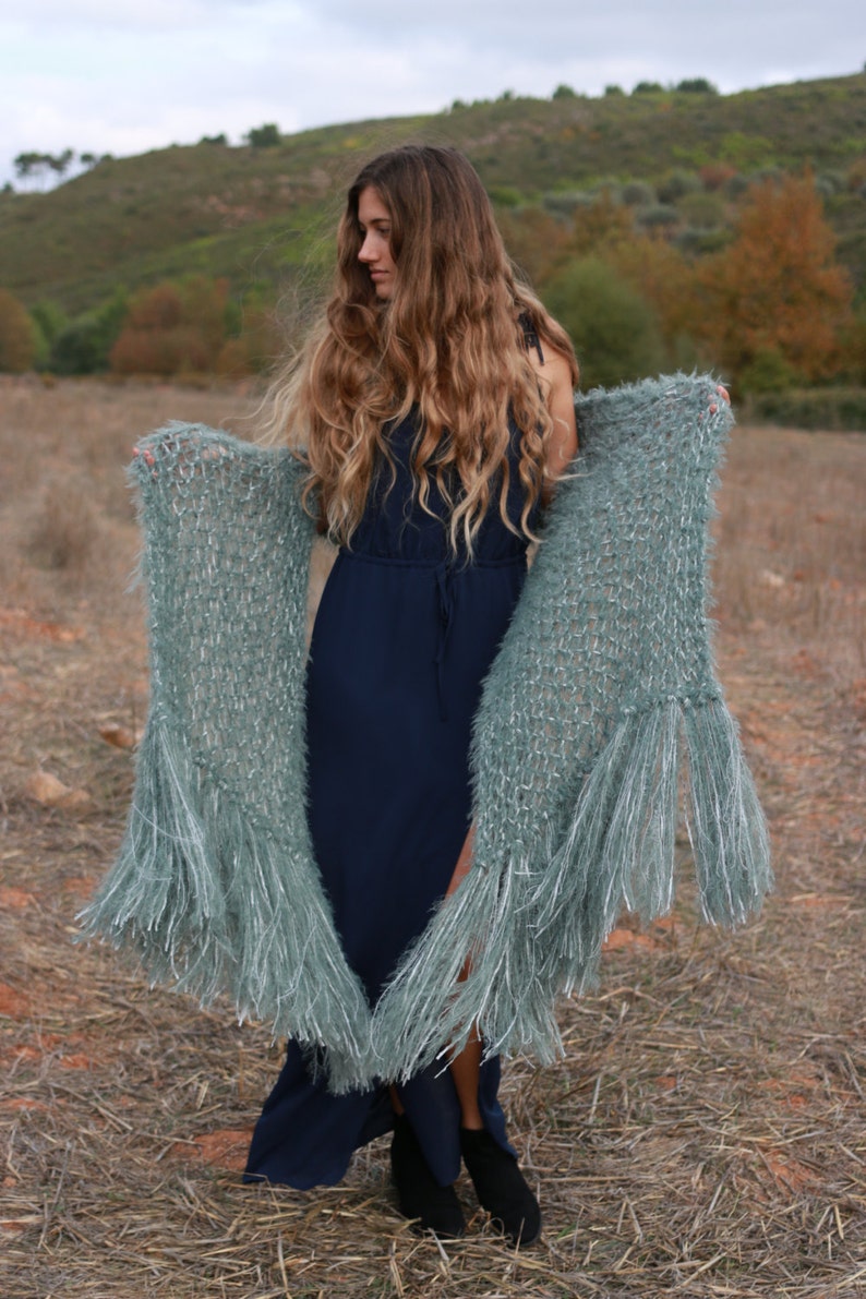 Puede incluir: Una mujer con un vestido azul marino largo y botas negras, sosteniendo un chal verde azulado claro con largas borlas. El chal est&aacute; hecho de un material suave y texturizado. El fondo presenta un campo y un cielo nublado.