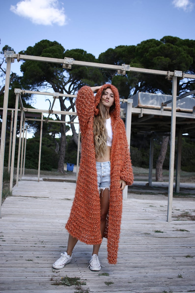 May include: A woman wearing a long, orange knitted cardigan with a hood. She is standing on a wooden platform with a wooden structure behind her.
