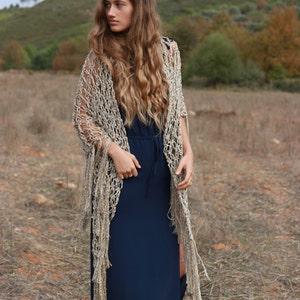 May include: A woman wearing a long, navy blue dress with a crocheted shawl in a beige and gold color. The shawl has long fringe and is draped over her shoulders. She is standing in a field of dry grass.