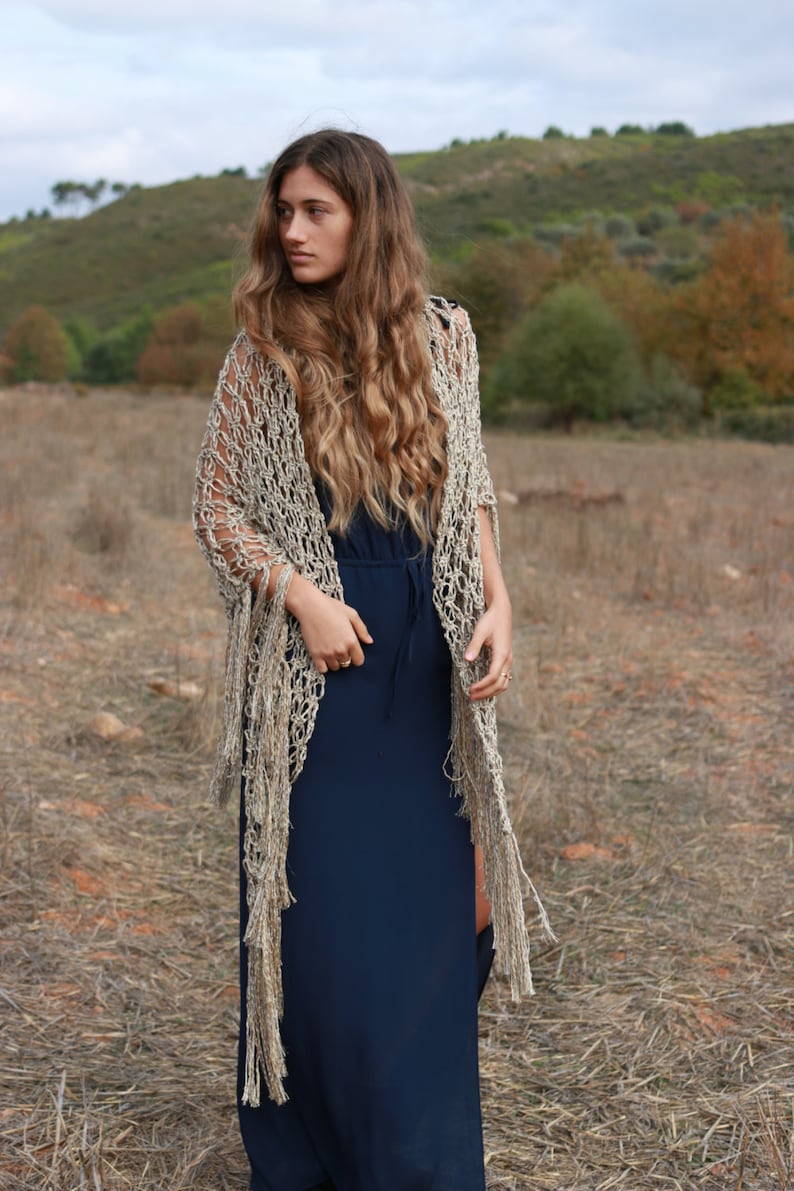 May include: A woman wearing a long, navy blue dress with a crocheted shawl in a beige and gold color. The shawl has long fringe and is draped over her shoulders. She is standing in a field of dry grass.