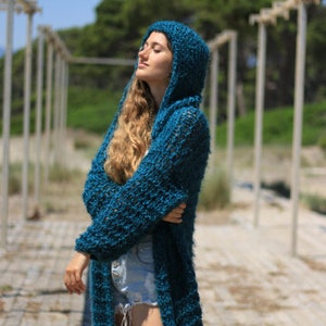 May include: A teal blue knitted cardigan with a hood. The cardigan is long and has a loose fit. The model is wearing the cardigan with a pair of denim shorts.