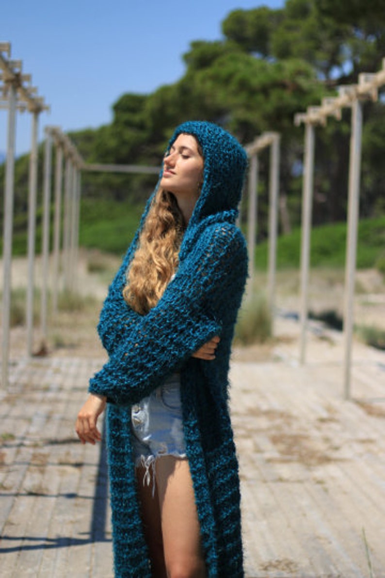 May include: A teal blue knitted cardigan with a hood. The cardigan is long and has a loose fit. The model is wearing the cardigan with a pair of denim shorts.