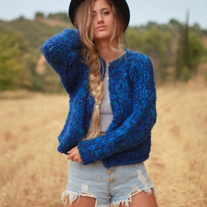 May include: A woman wearing a blue and green knit cardigan with a black hat and denim shorts.