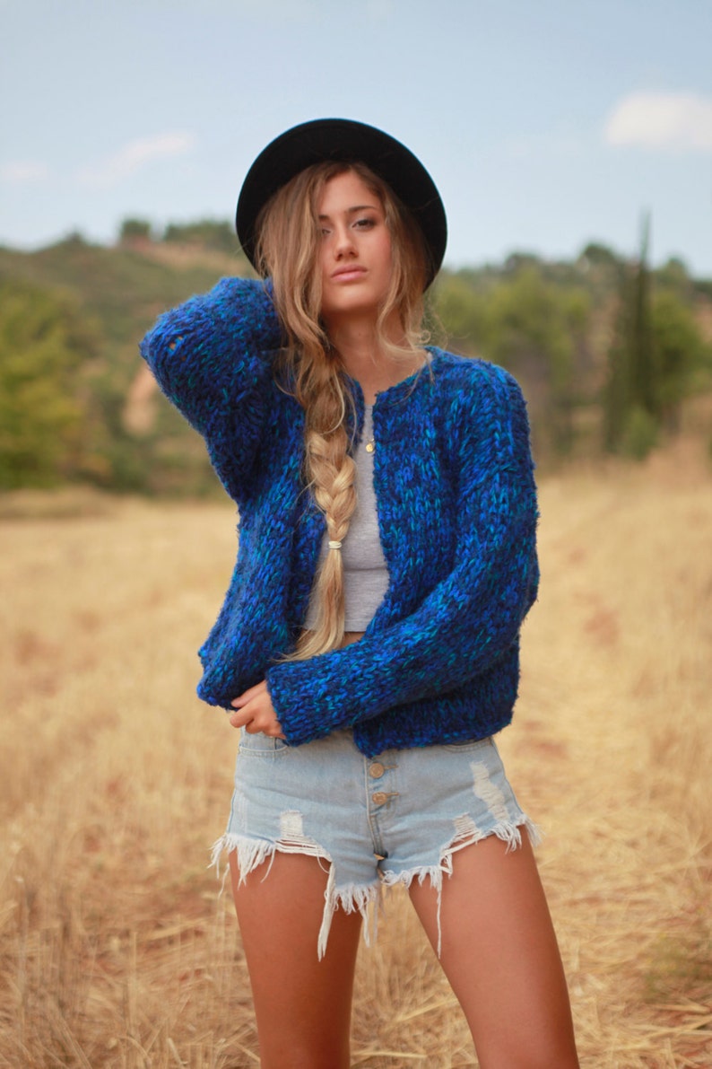 May include: A woman wearing a blue and green knit cardigan with a black hat and denim shorts.