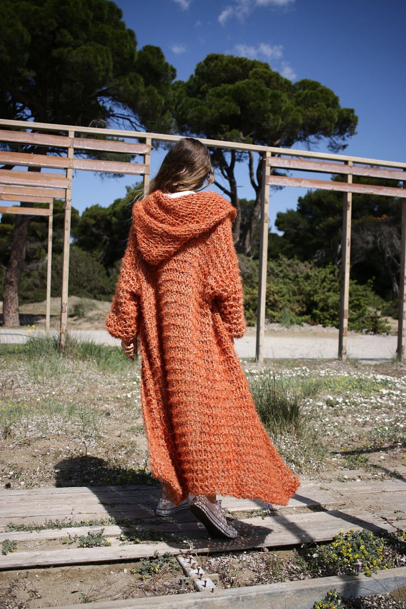 May include: A long, orange knitted cardigan with a hood. The cardigan is made of a soft, fluffy yarn and has a relaxed fit.