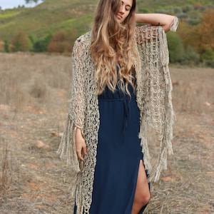 May include: A woman wearing a long, navy blue dress with a side slit. She is wearing a crocheted shawl with fringe that is a light beige color. The shawl is draped over her shoulders and hangs down to her ankles. She is standing in a field of dry grass.