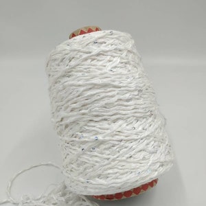 May include: A spool of white yarn with silver sequins woven throughout. The yarn is wound around a cardboard spool with red and white markings.