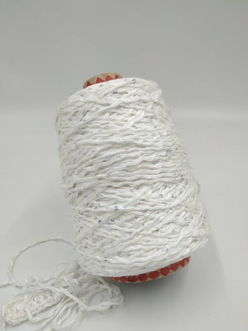 May include: A spool of white yarn with silver sequins woven throughout. The yarn is wound around a cardboard spool with red and white markings.