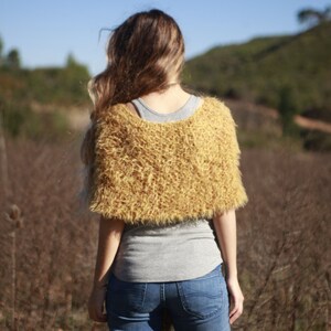 May include: A woman wearing a fuzzy, mustard yellow knitted shawl over a gray shirt and blue jeans. She is standing in a field of dry grass.