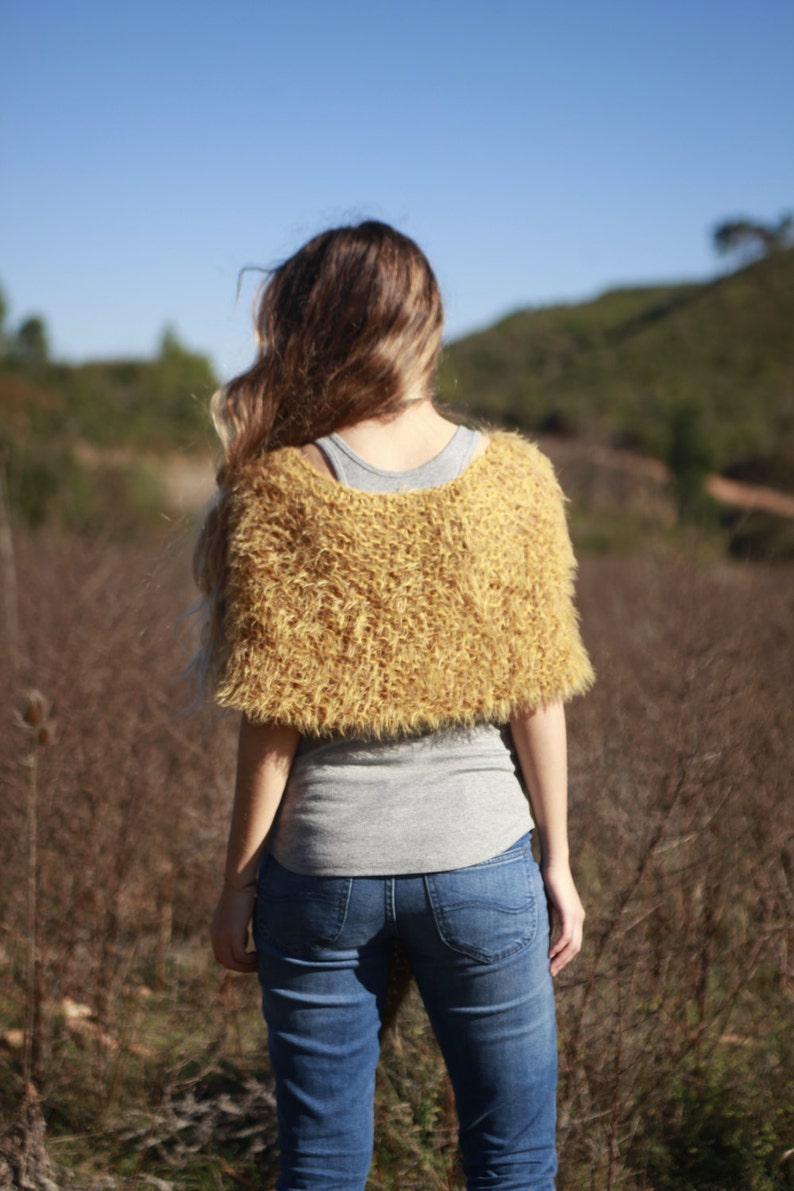 May include: A woman wearing a fuzzy, mustard yellow knitted shawl over a gray shirt and blue jeans. She is standing in a field of dry grass.