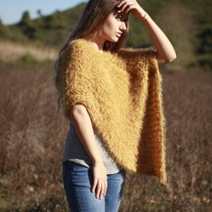 May include: A woman wearing a fuzzy, mustard yellow poncho with a gray shirt and blue jeans. She is standing in a field of tall grass.