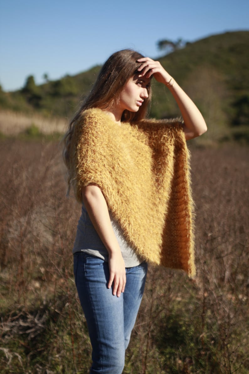 May include: A woman wearing a fuzzy, mustard yellow poncho with a gray shirt and blue jeans. She is standing in a field of tall grass.