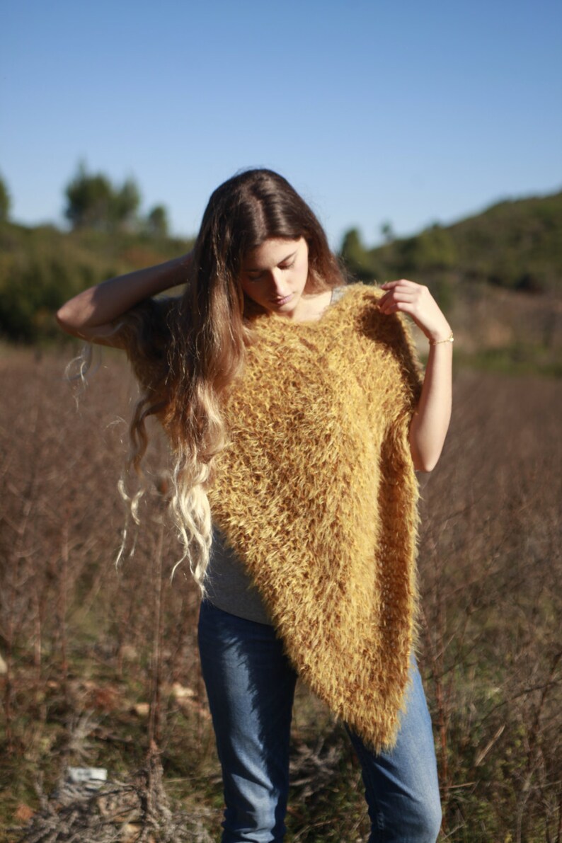 May include: A woman wearing a long, fuzzy, mustard yellow poncho with long brown hair. She is standing in a field of tall grass.