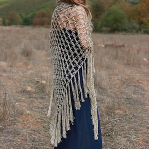 May include: A woman wearing a long, navy blue dress with a crocheted shawl in a beige color. The shawl has fringe detailing and is worn over the shoulders. The woman is standing in a field of dry grass.