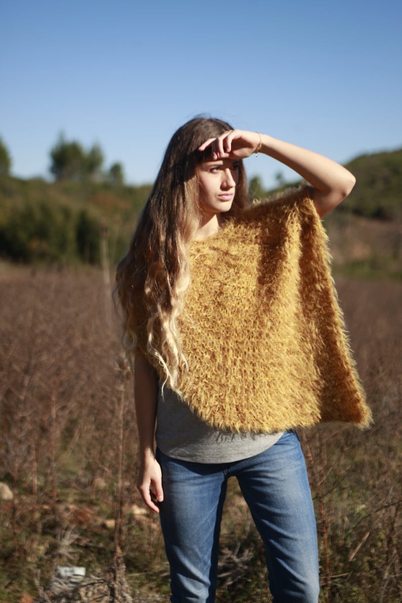 May include: A woman wearing a fuzzy, mustard yellow poncho with a gray shirt and blue jeans. She is standing in a field of brown grass.