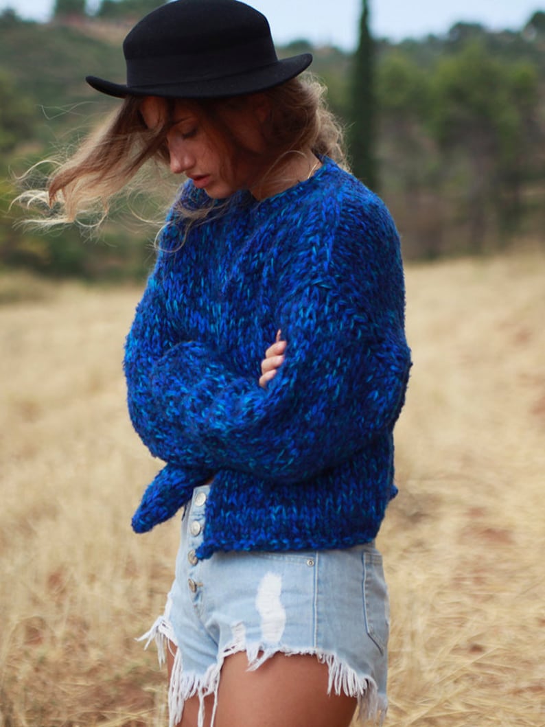 May include: A woman wearing a blue fuzzy sweater with a black fedora hat and light blue denim shorts with frayed edges.