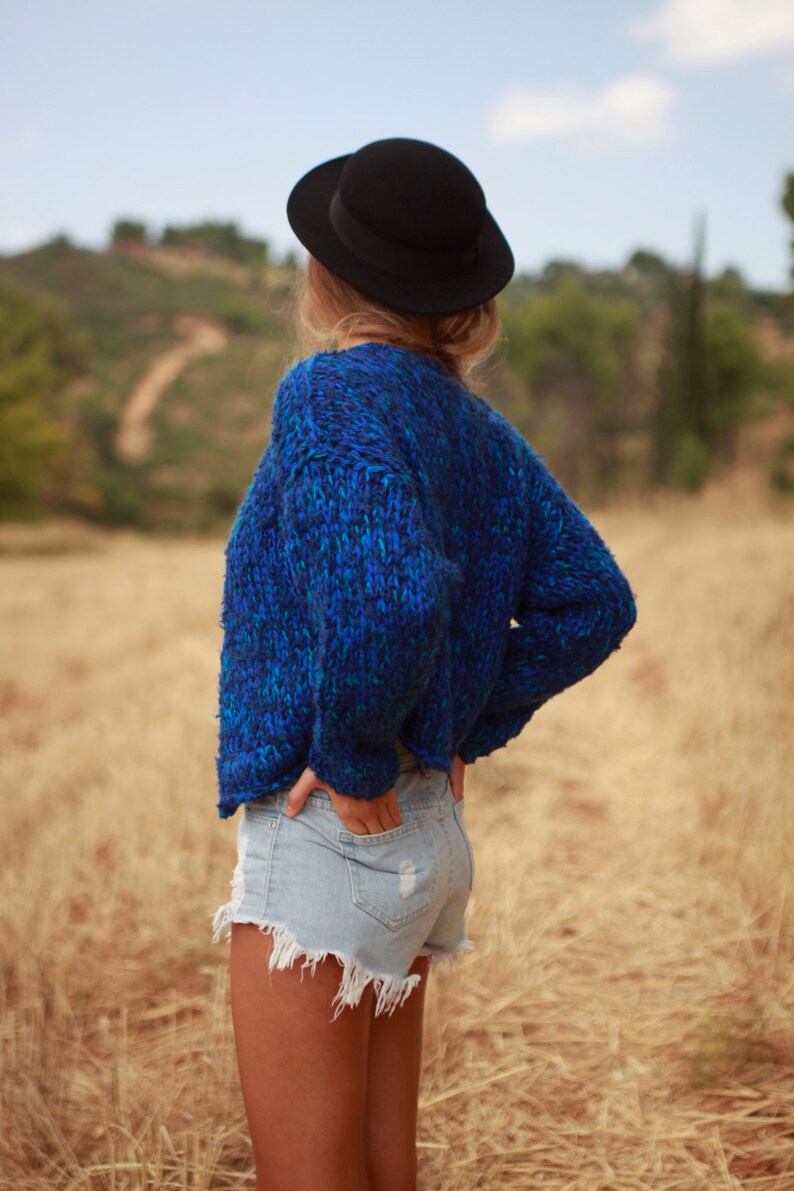 May include: A woman wearing a blue knitted sweater and denim shorts stands in a field of tall grass. She is wearing a black hat and has her back to the camera.